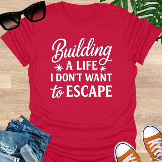 White script with block text: “Building a life I don’t want to escape” on red unisex tee.