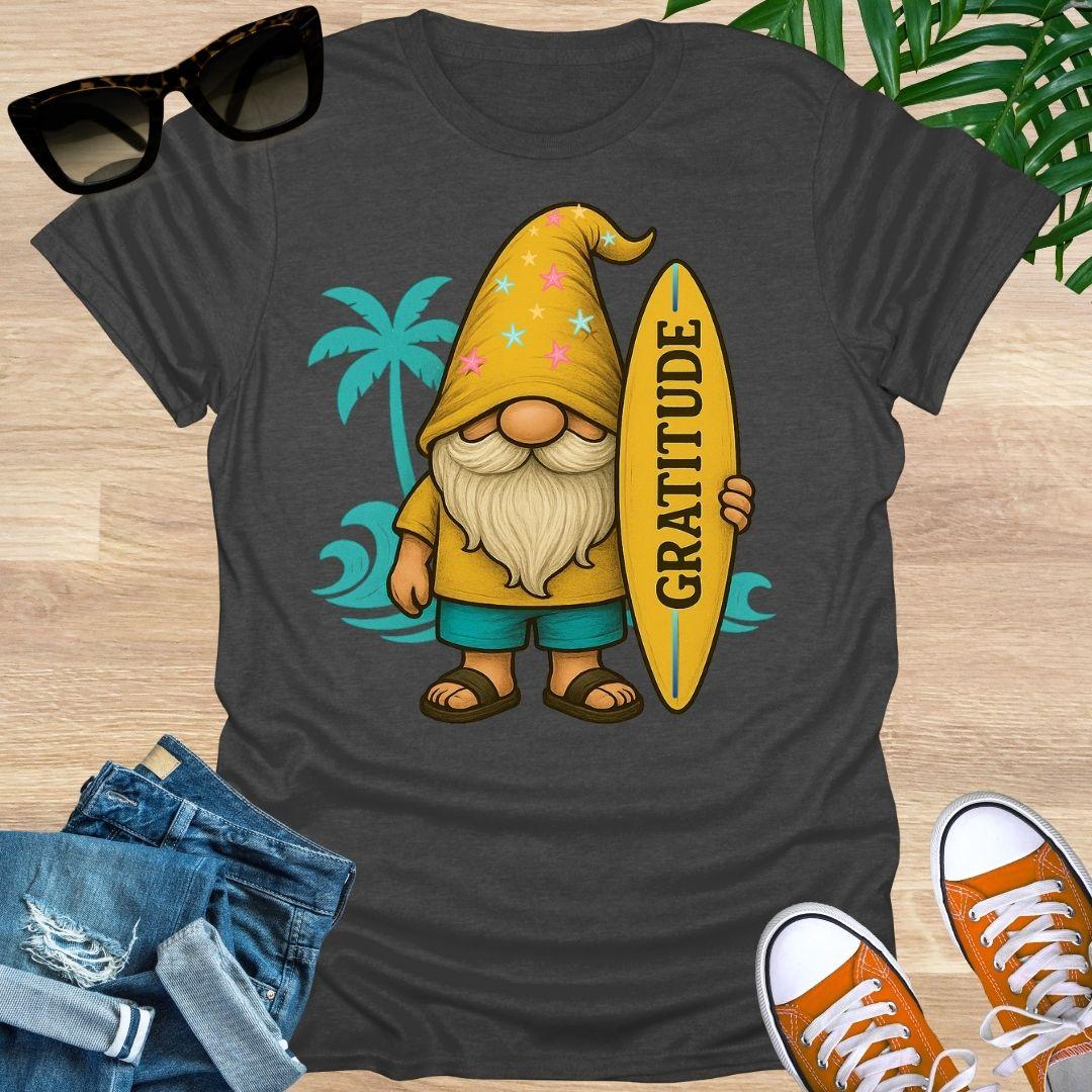 Heather Gray t-shirt with a cartoon gnome holding a surfboard with 'Gratitude' on it, sunglasses, jeans, and shoes on a wooden surface.