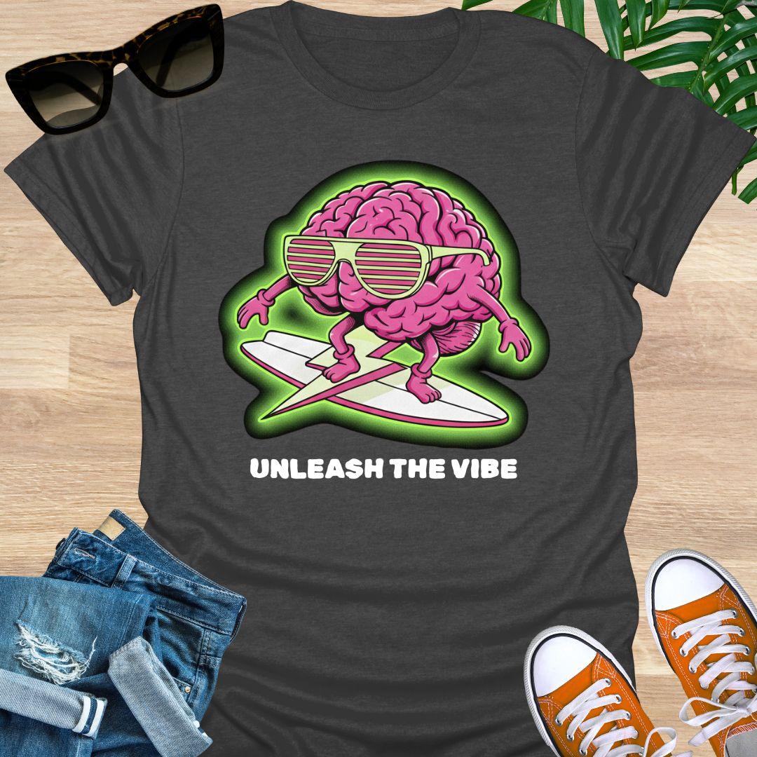 Pink cartoon brain wearing sunglasses surfing on a board with green glow and the words “Unleash the Vibe” on a heather gray t-shirt