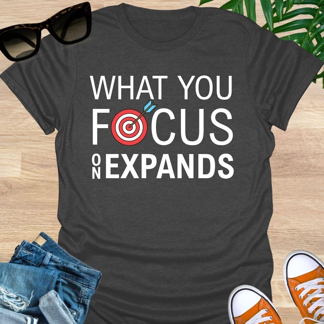 “What You Focus On Expands” text with red bullseye and arrow on a heather gray unisex tee. 