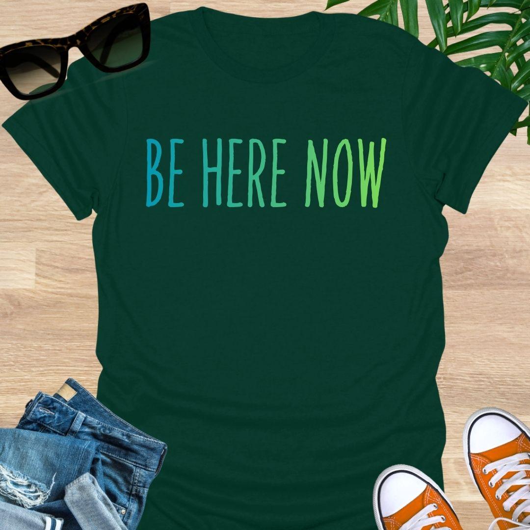 Blue-green gradient “Be Here Now” text on forest green unisex mindfulness tee; reminder to breathe and be present.