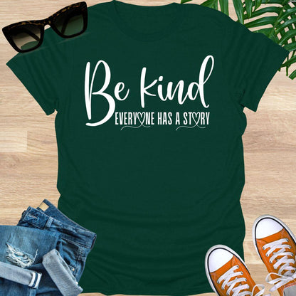 “Be Kind—Everyone Has a Story” in white script with heart loops on a forest green unisex tee.