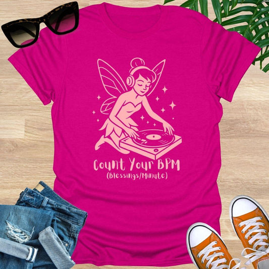 Pink t-shirt with a graphic of a fairy DJ and text 'Count Your BPM' on a wooden surface with sunglasses, jeans, and sneakers.