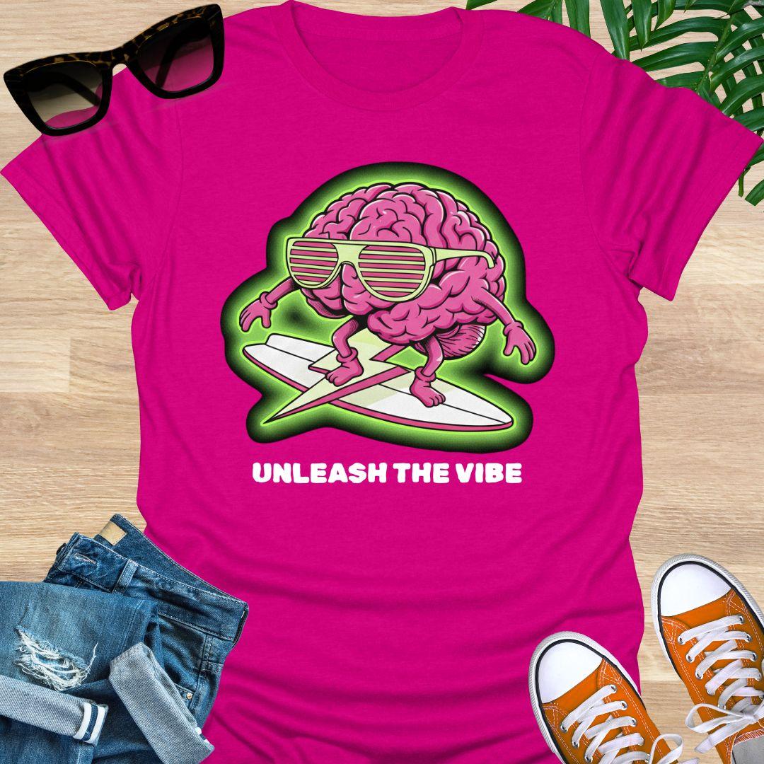 Pink cartoon brain wearing sunglasses surfing on a board with green glow and the words “Unleash the Vibe” on a hot pink t-shirt