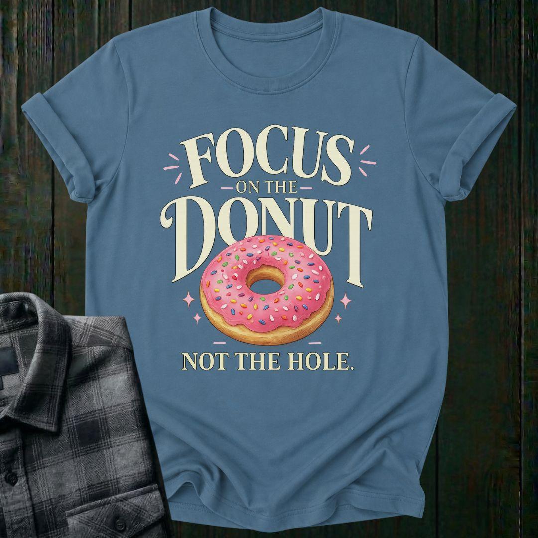Typography design featuring the phrase "Focus on the Donut, Not the Hole" with a pink sprinkled donut, symbolizing gratitude, perspective, and focusing on life's blessings on a light blue tee