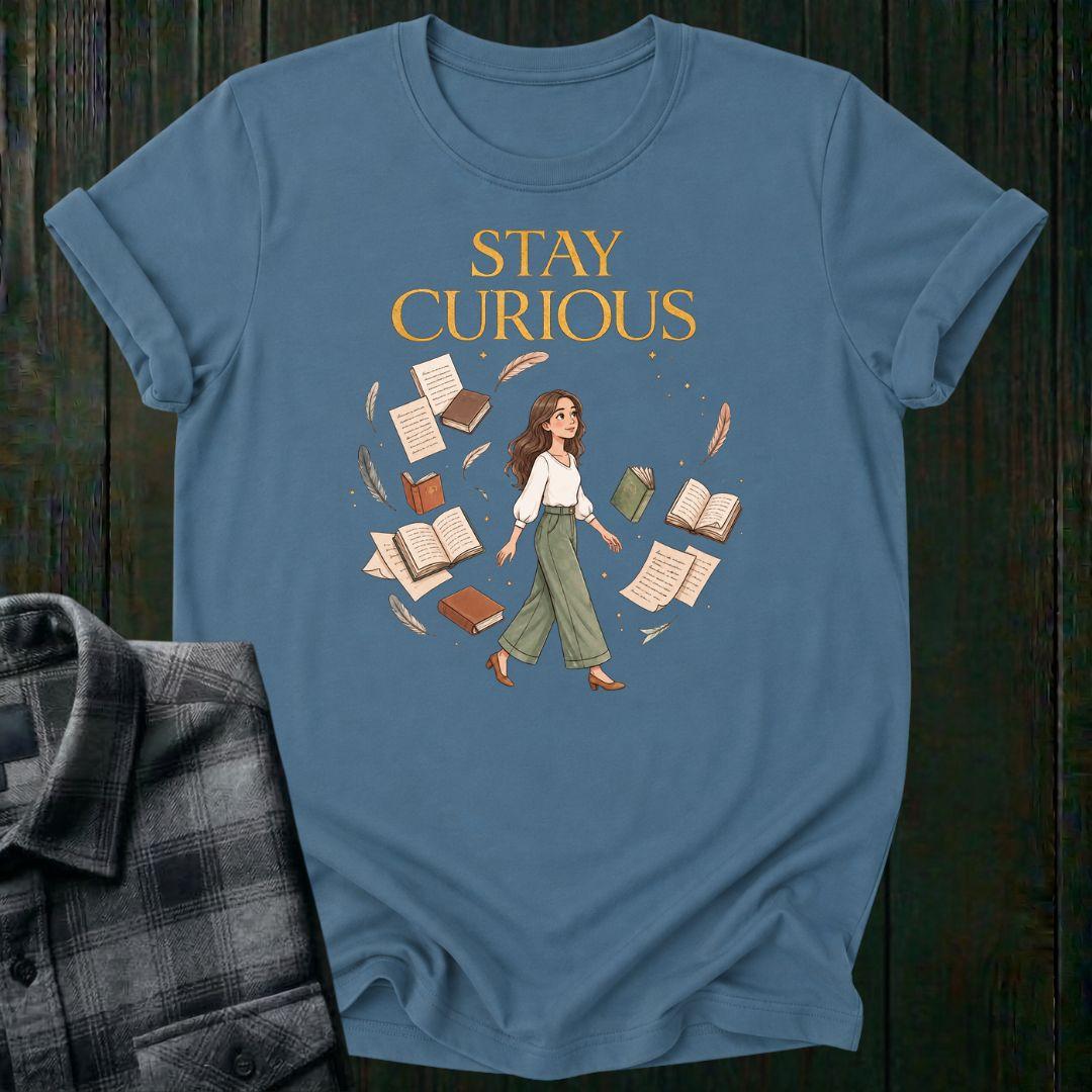 Illustrated woman walking among floating books, journals, feathers, and stars beneath the words "Stay Curious," symbolizing lifelong learning, creativity, and wonder on a light blue tee.