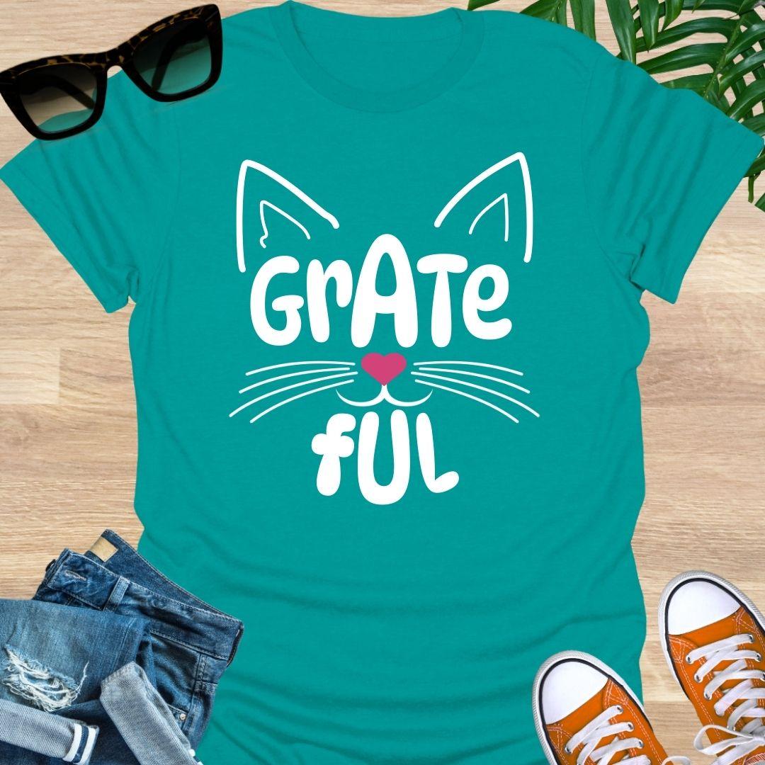 Teal unisex graphic t-shirt with cat ears, whiskers, and a pink heart nose forming the word GRATEFUL
