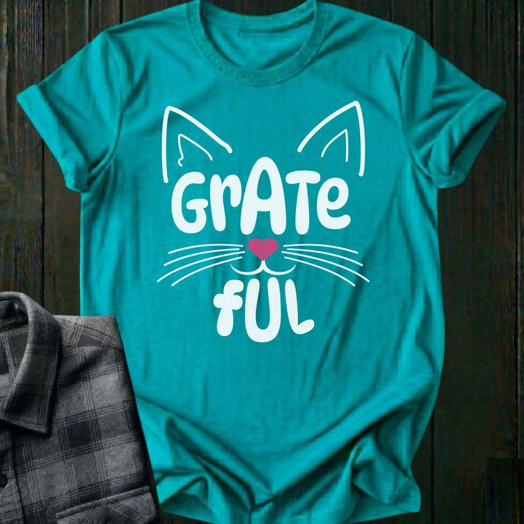 Turquoise t-shirt with a cat-themed design and 'Grate Ful' text on a wooden surface.