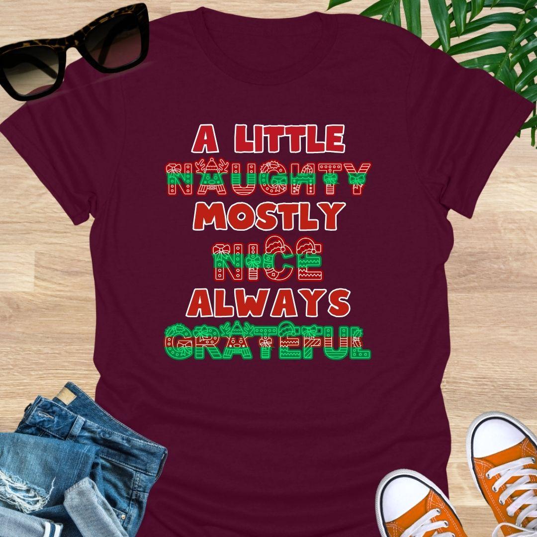 Red-green holiday letters spelling “A Little Naughty, Mostly Nice, Always Grateful” with bows, wreaths, and reindeer on a maroon unisex tee.