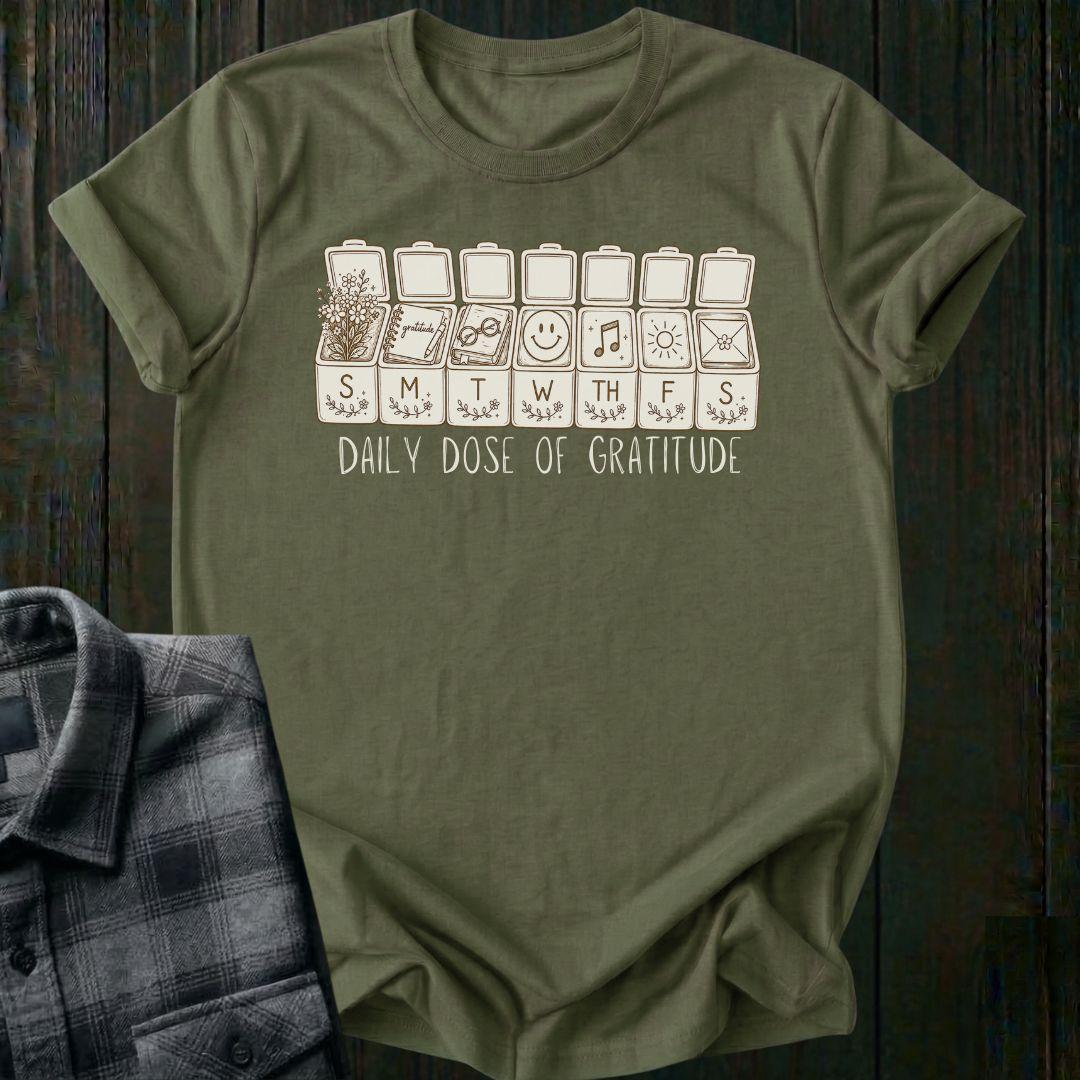 Vintage-inspired weekly pill organizer graphic featuring gratitude-themed icons for each day of the week, including flowers, journaling, reading, smiling, music, sunshine, and connection, with the phrase Daily Dose of Gratitude on a military green tee