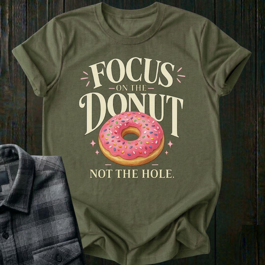 Typography design featuring the phrase "Focus on the Donut, Not the Hole" with a pink sprinkled donut, symbolizing gratitude, perspective, and focusing on life's blessings on a military green tee