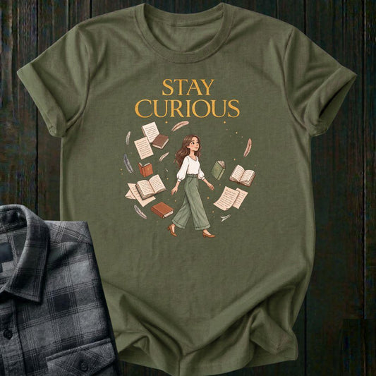 Illustrated woman walking among floating books, journals, feathers, and stars beneath the words "Stay Curious," symbolizing lifelong learning, creativity, and wonder on a military green tee.