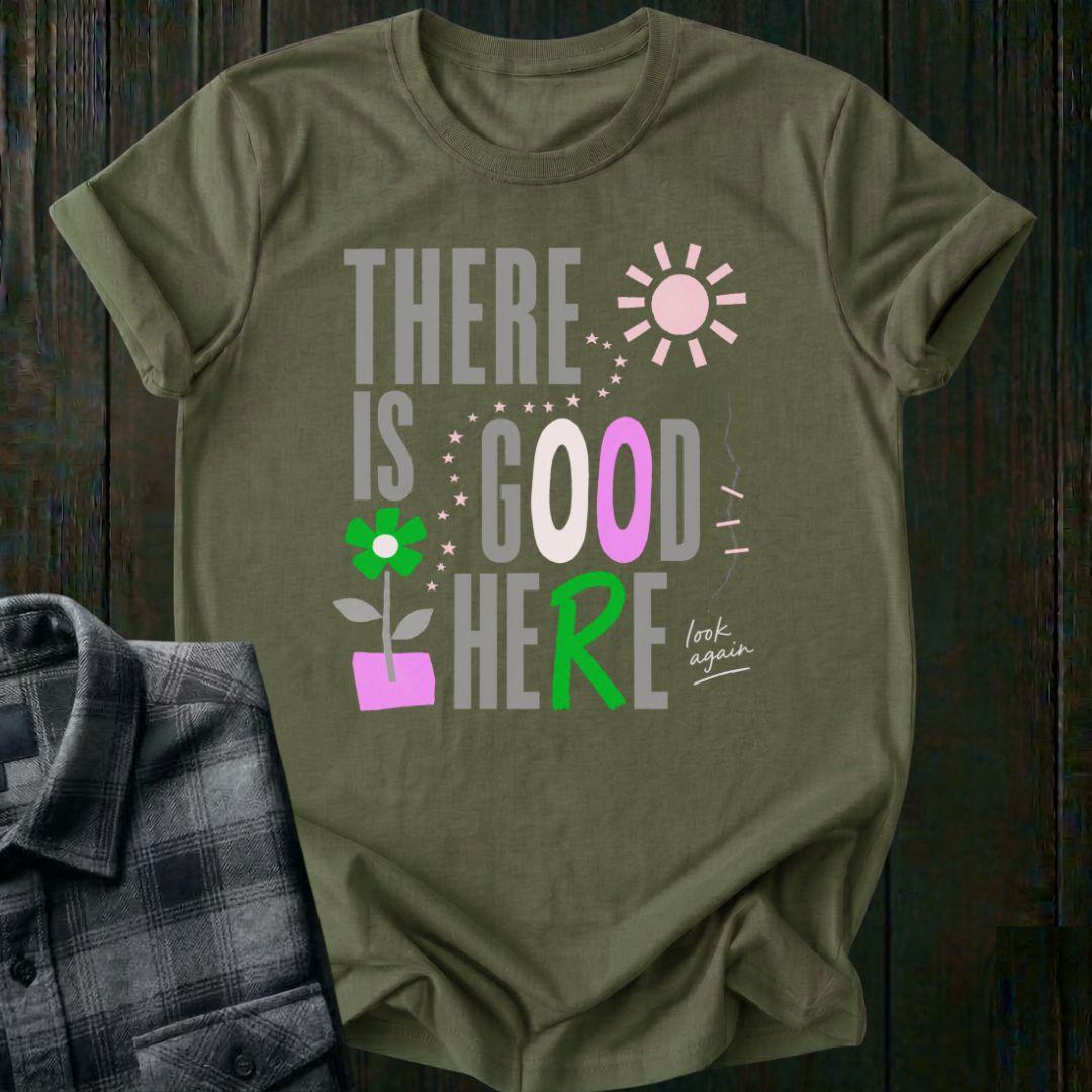 Colorful typography design featuring the phrase "There Is Good Here" with flowers, stars, sunshine, and the handwritten words "look again" on a military green tee.
