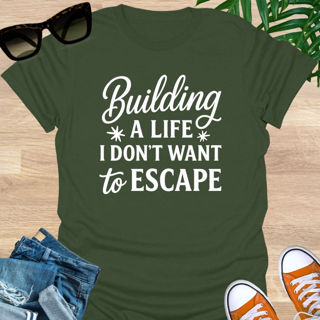White script with block text: “Building a life I don’t want to escape” on military green unisex tee.