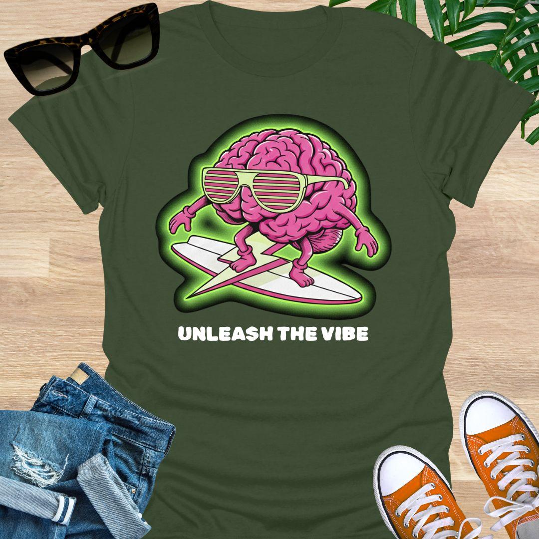 Pink cartoon brain wearing sunglasses surfing on a board with green glow and the words “Unleash the Vibe” on a military green t-shirt