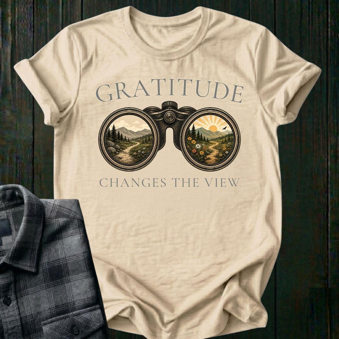 Vintage binoculars graphic showing two landscapes, one ordinary and one bright and colorful, with the phrase "Gratitude Changes the View" on a beige tee