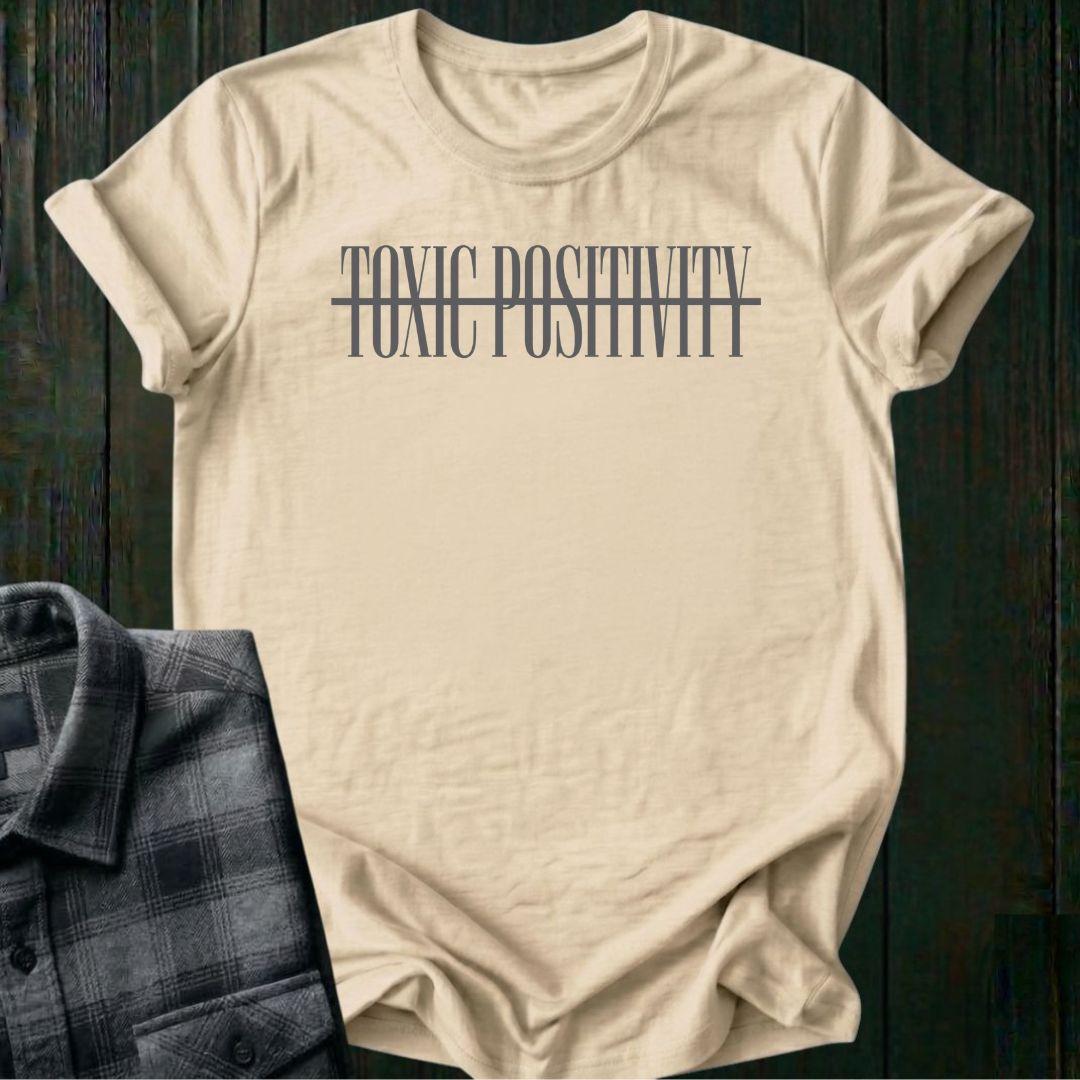 Natural tee with minimalist typography design featuring the phrase "No More Toxic Positivity" in bold modern lettering with a horizontal line running through the center of the text.