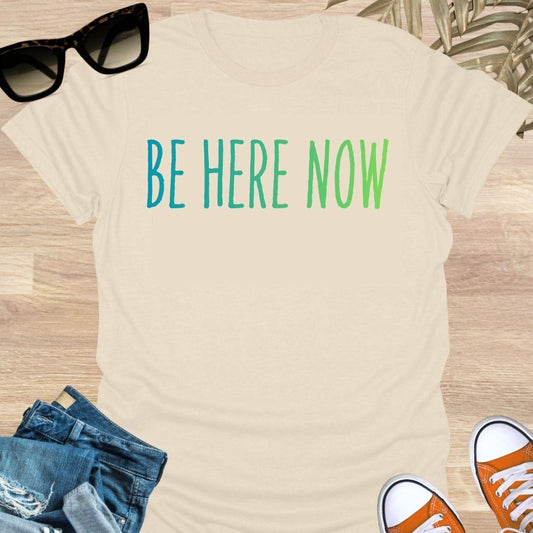 Blue-green gradient “Be Here Now” text on beige unisex mindfulness tee; reminder to breathe and be present.
