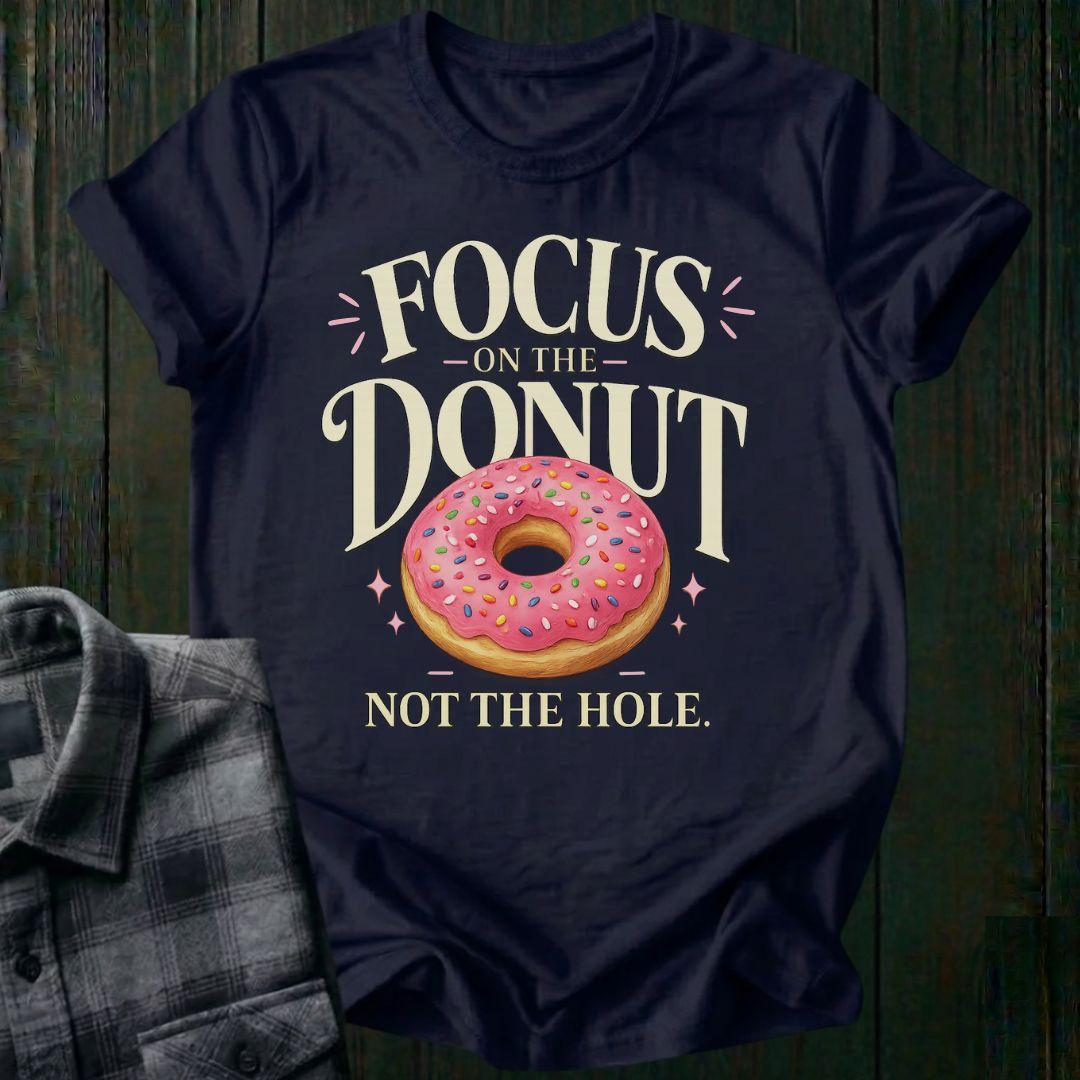 Typography design featuring the phrase "Focus on the Donut, Not the Hole" with a pink sprinkled donut, symbolizing gratitude, perspective, and focusing on life's blessings on a navy tee