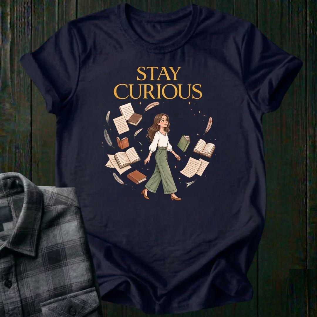 Illustrated woman walking among floating books, journals, feathers, and stars beneath the words "Stay Curious," symbolizing lifelong learning, creativity, and wonder on a navy tee.