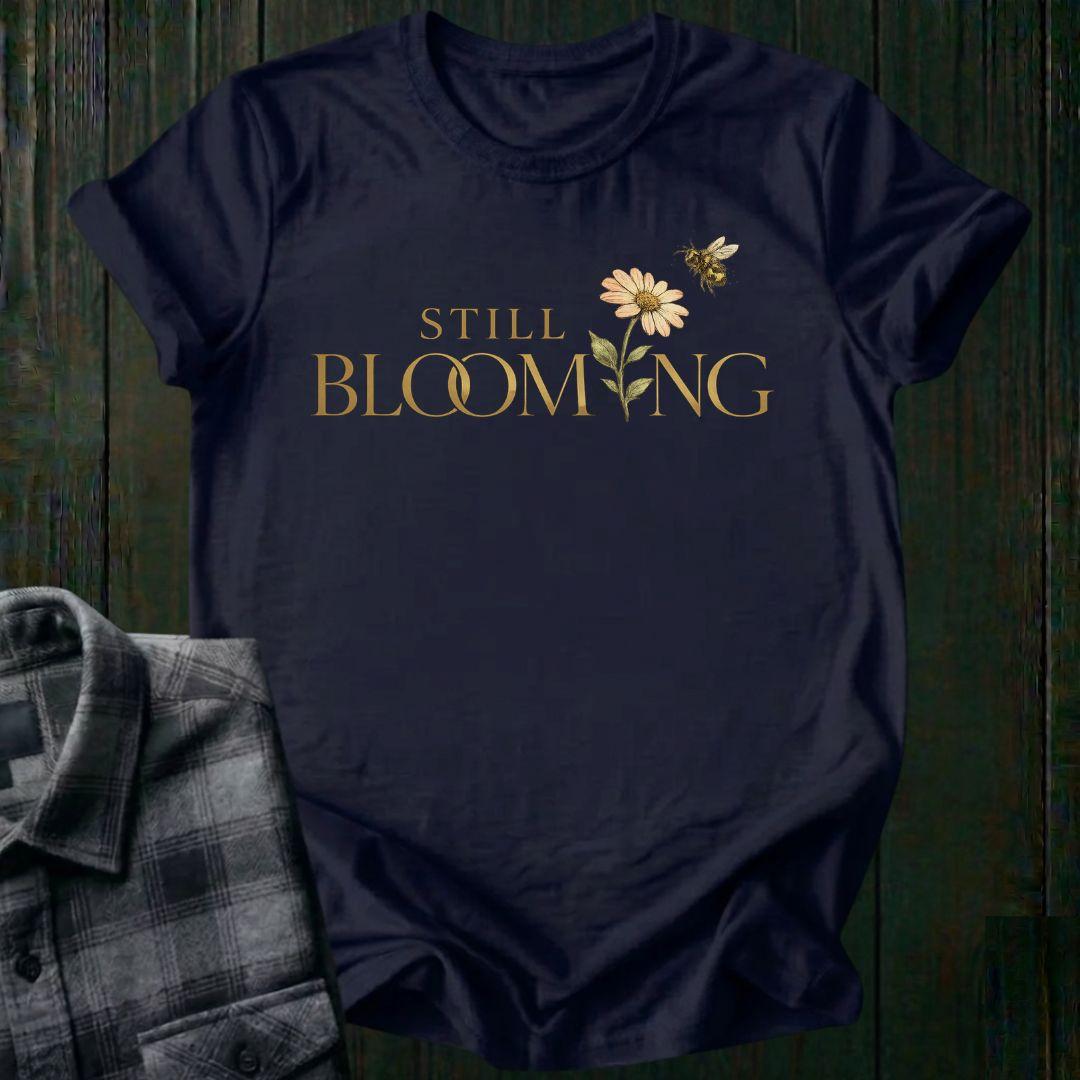 Elegant typography design featuring the phrase "Still Blooming" with a blooming flower growing through the lettering and a bee flying overhead on a navy shirt