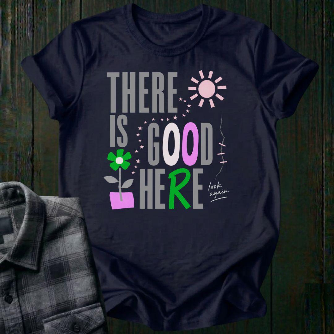 Colorful typography design featuring the phrase "There Is Good Here" with flowers, stars, sunshine, and the handwritten words "look again" on a navy tee.
