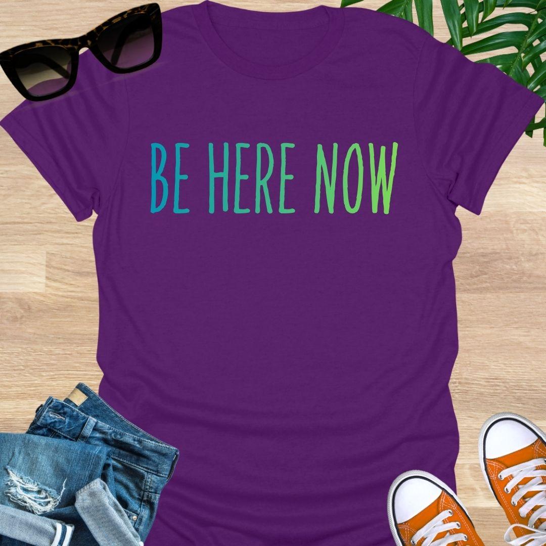 Blue-green gradient “Be Here Now” text on purple unisex mindfulness tee; reminder to breathe and be present.