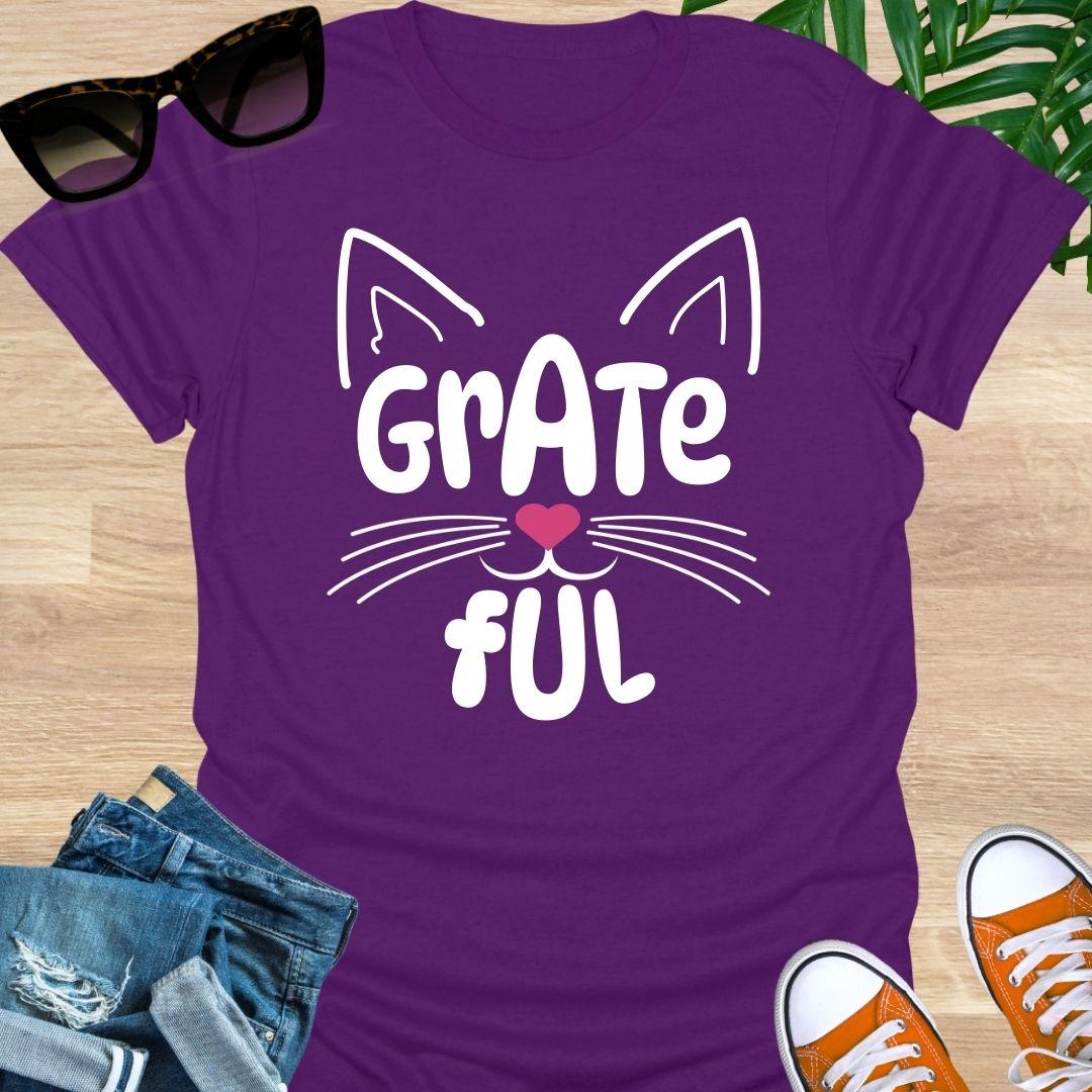 Purple unisex graphic t-shirt with cat ears, whiskers, and a pink heart nose forming the word GRATEFUL