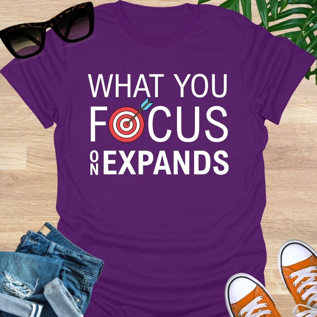  “What You Focus On Expands” text with red bullseye and arrow on a purple unisex tee. 