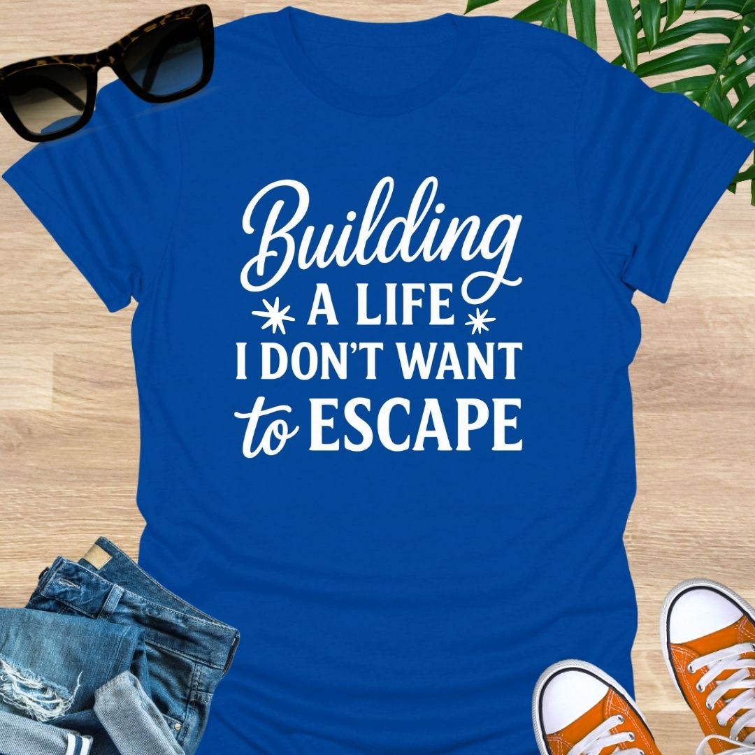 White script with block text: “Building a life I don’t want to escape” on blue unisex tee.