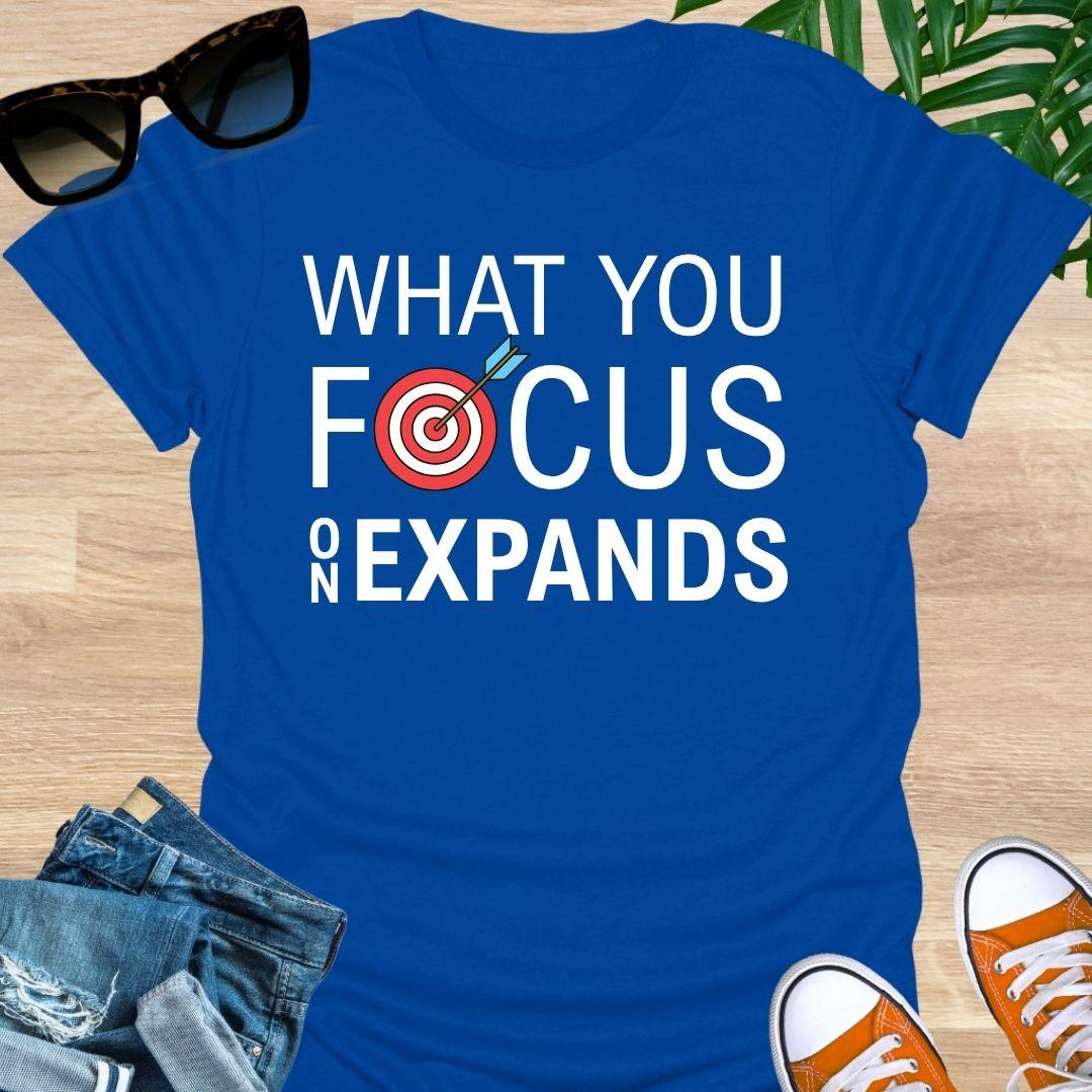  “What You Focus On Expands” text with red bullseye and arrow on a blue unisex tee. 