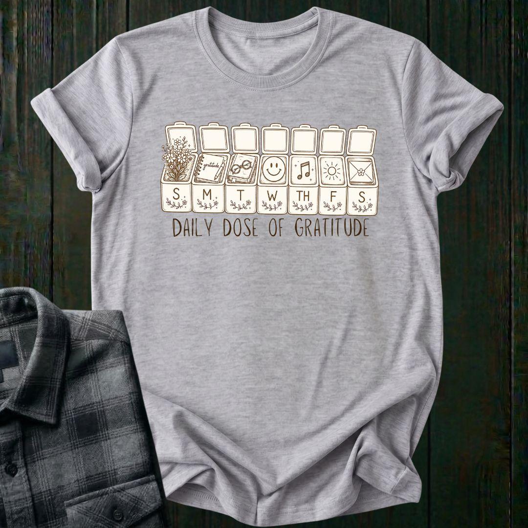 Vintage-inspired weekly pill organizer graphic featuring gratitude-themed icons for each day of the week, including flowers, journaling, reading, smiling, music, sunshine, and connection, with the phrase Daily Dose of Gratitude on a sports gray tee