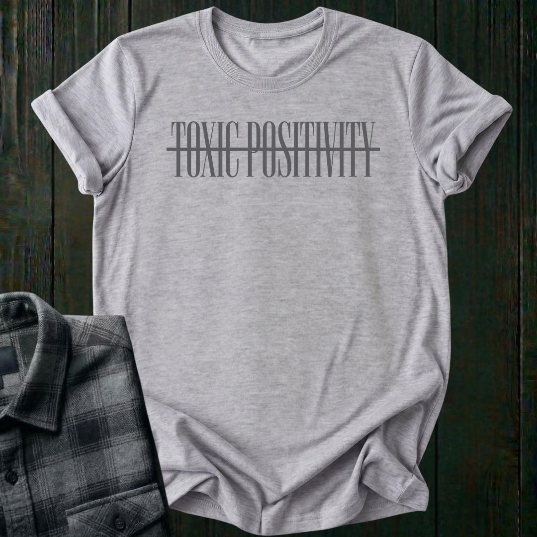 light gray tee with minimalist typography design featuring the phrase "No More Toxic Positivity" in bold modern lettering with a horizontal line running through the center of the text.