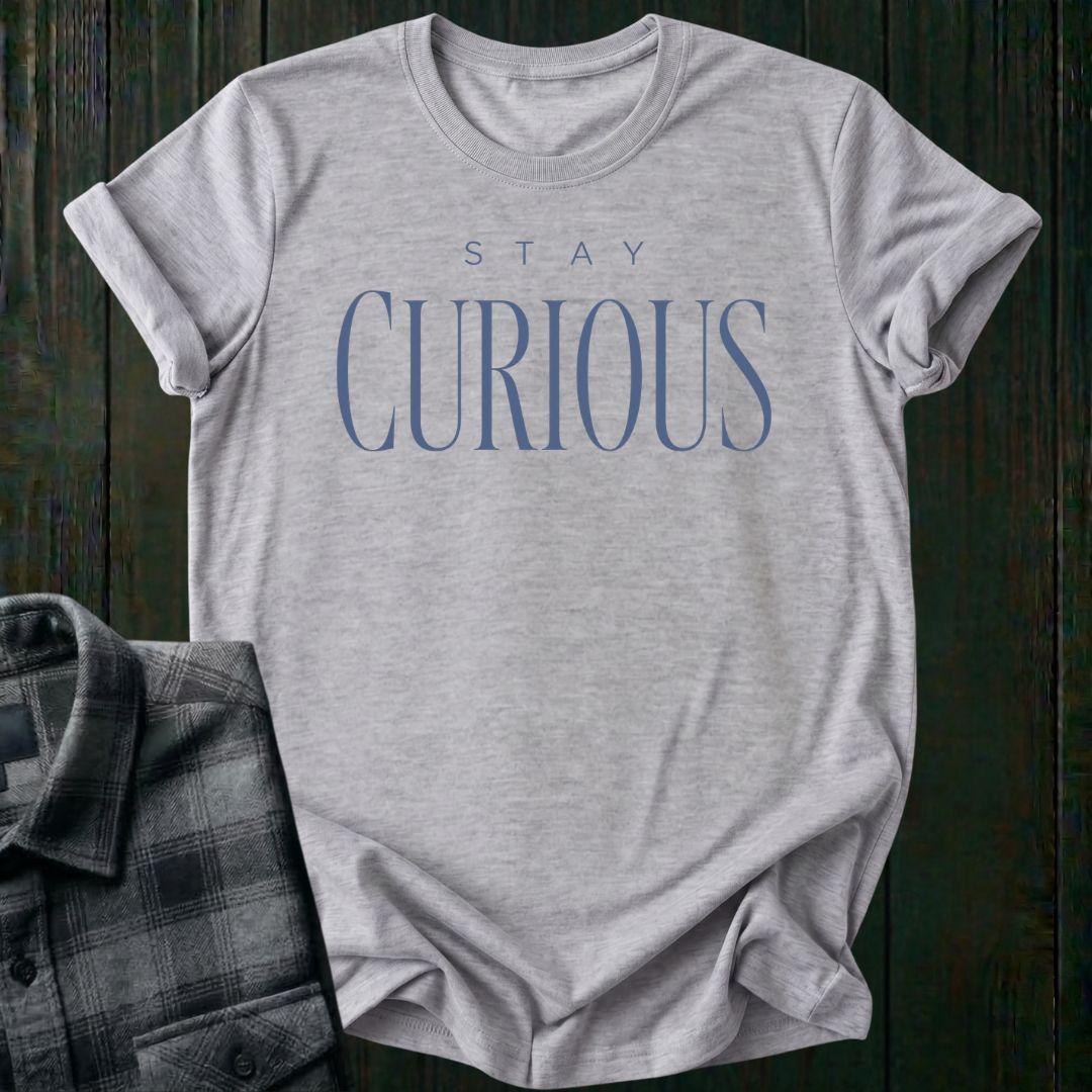 Elegant minimalist typography design featuring the phrase "Stay Curious" in refined serif lettering on a light gray tee.